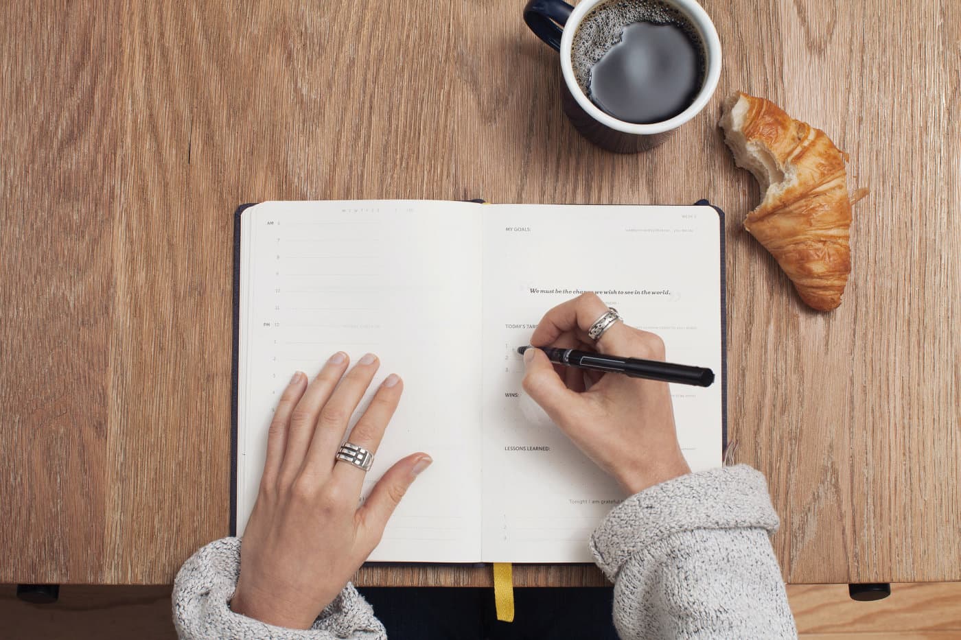 A table with a notebook, a coffee mug and a croissant from above. A women is taking notes.